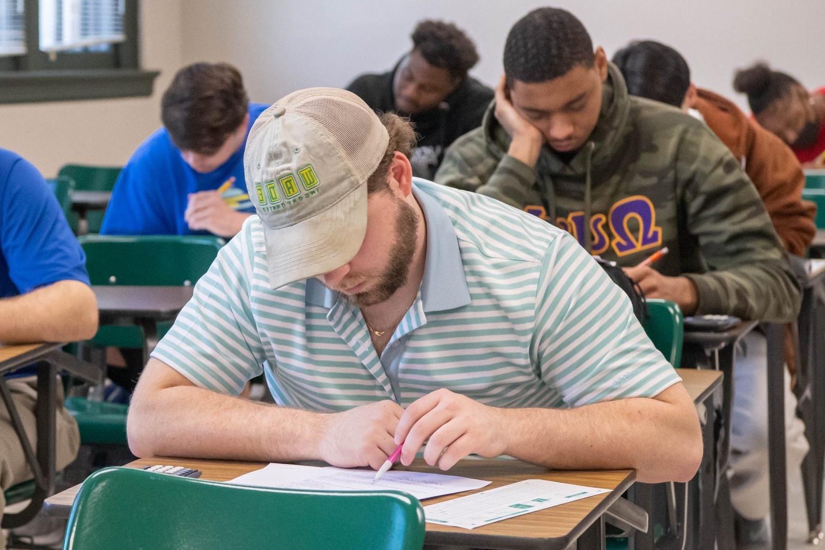 Student sitting at a desk completing assignment during class, with another six students behind him also at desks completing the assignment.